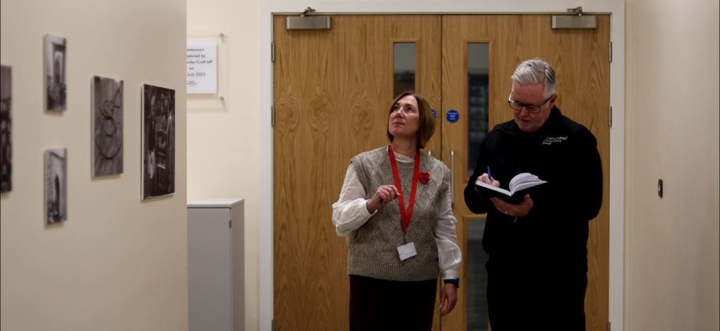 a women looking up in a school building with a man stood beside her writing notes down